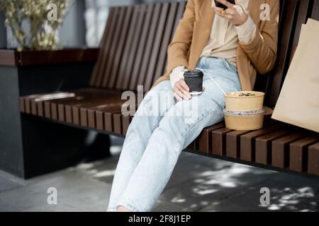 Pretty woman have outdoor lunch near office building while sitting on the bench and using phone. Healthy meal and coffee for takeaway for workers.  Stock Photo