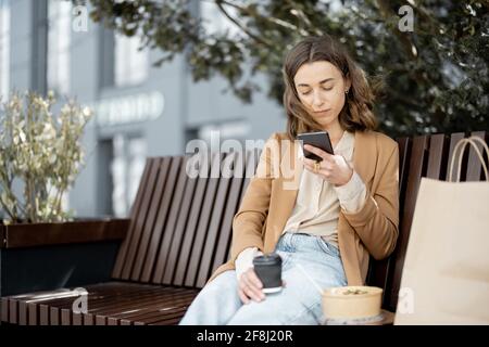 Pretty woman have outdoor lunch near office building while sitting on the bench and using phone. Healthy meal and coffee for takeaway for workers.  Stock Photo