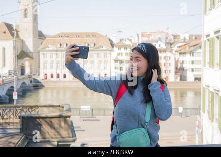 Asian woman backpacker touring in Europe - young happy and pretty Japanese girl with student backpack taking selfie with mobile phone during holidays Stock Photo