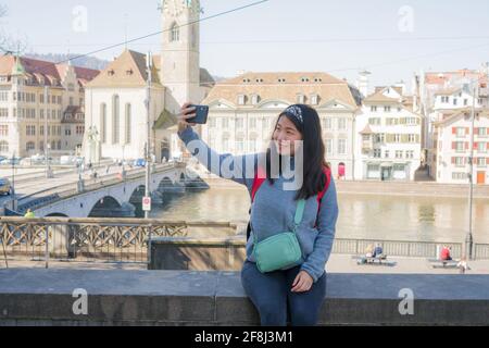 Asian woman backpacker touring in Europe - young happy and pretty Korean girl with student backpack taking selfie with mobile phone during holidays in Stock Photo