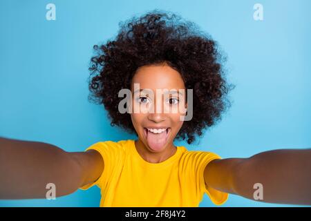 Photo funny dark skin brown haired little girl wear yellow t-shirt take selfie fooling face isolated on blue color background Stock Photo