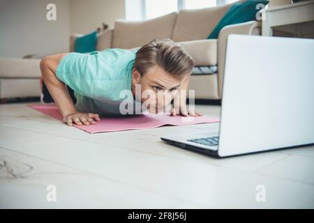 Caucasian man with blonde hair is doing push-ups on the floor while using a laptop