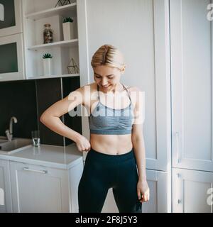 Caucasian woman with sporty body is smiling in the kitchen wearing sportswear during a fitness warming up Stock Photo
