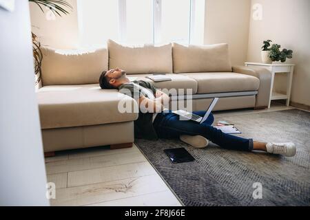 Sleepy caucasian man is working on the floor on a laptop with some documents while leaning on a couch and resting Stock Photo