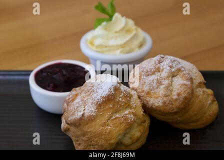 Hot scones sprinkled with powdered sugar and accompanied by whipped butter cream and red jam on rustic dark ceramic plate on light wood table. Stock Photo