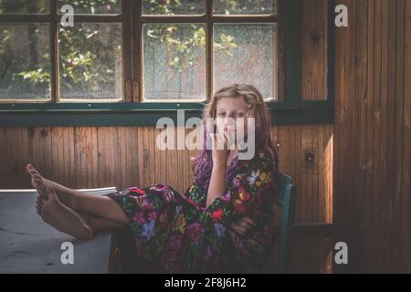 adorable girl with long blond hair sitting on chair behind the window and relaxing Stock Photo