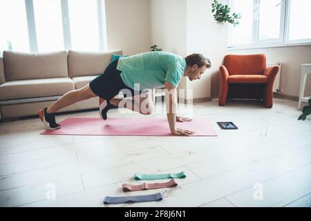 Blonde man is using a tablet while doing fitness exercises at home on a yoga mat Stock Photo