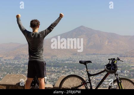 Young man with arms up next to bike with views of Santiago city on sunny day. Triumphant cyclist on top of mountain in capital of Chile Stock Photo