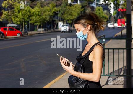 Middle aged woman with face mask on looking at cellphone while waiting on bus stop on sunny day in Santiago city, Chile. Slim lady using mobile phone