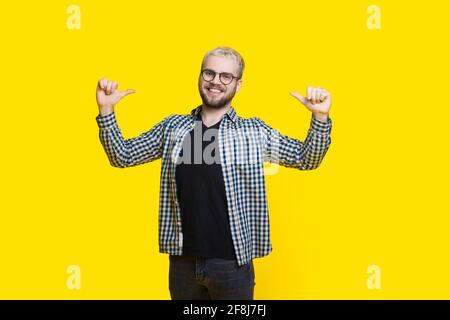 Self proud man is gesturing at himself smiling on a yellow studio wall through glasses wearing a beard Stock Photo