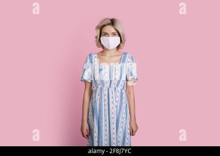 Blonde woman in a long summer dress is wearing a medical mask on a pink studio wall Stock Photo