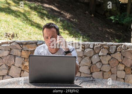 A man working at home outdoors using a laptop and talking on the cell Stock Photo