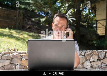 A man working at home outdoors using a laptop and talking on the cell Stock Photo