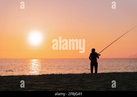 Silhouette of man fishing in waves on beach at sunset Stock Photo