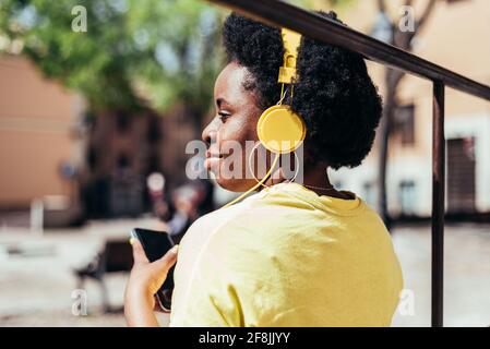 Rear view of a black girl with afro hair and hoop earrings listening to music with her cell phone and yellow headphones in an urban space in the city. Stock Photo