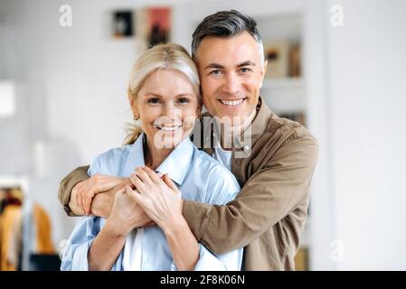 Close up portrait of lovely caucasian marriage couple, happy husband and wife dressed in casual stylish clothes, standing at home, hugging, looking smiling directly into the camera Stock Photo