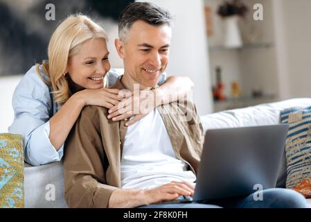 A middle aged married happy caucasian couple at home on the couch is using laptop. Joyful husband and wife browsing internet, social media, chatting with friends, do online shopping for home, smiling Stock Photo