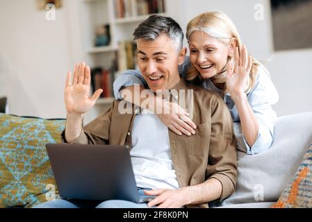 Happy caucasian joyful spouses spend time at home on a sofa, use laptop for online communication with friends or family by video conference, greeting by waving hands, smiling. Video call concept Stock Photo
