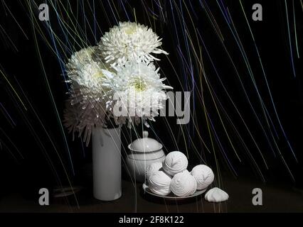 Modern still life with white flowers in a white vase, white dishes and marshmallow in a white plate, with unusual luminous lines on a black background Stock Photo