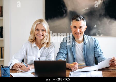 Intelligent businessman and business lady. Two successful partners, sit at the workplace, working on a new project, using a laptop, looking through documents, looking at the camera smiling friendly Stock Photo