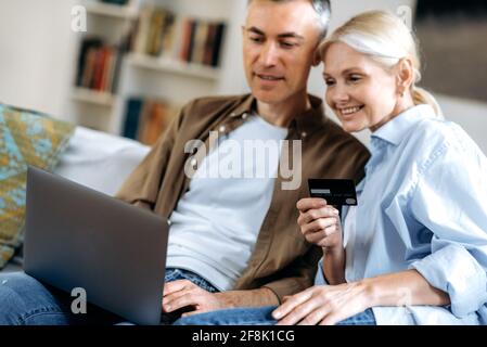 Defocused happy caucasian smiling couple in love, joyful husband and wife, using laptop and credit card for online payment of goods, clothes or delivery, online shopping, e-commerce concept Stock Photo