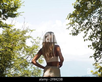 A tanned blonde stands near green trees looking into light cloudy haze and the blue sky Stock Photo