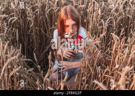 Portrait of young, teen, ginger girl with freckles in the wheat field with closed eyes, holding red bouquet. Copy space Stock Photo
