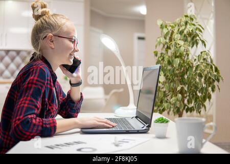 Blonde girl with glasses work on a laptop and talk on the phone. Home-school concept Stock Photo