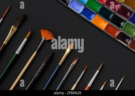 Different shape and size paintbrushes, set of watercolor paints on black paper sheet, top view, flat lay. Stock Photo