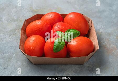Tomatoes and basil in drops of water, close-up in a cardboard box on a gray background. Side view, close-up. Stock Photo