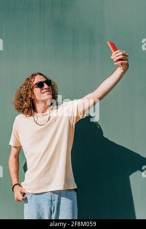 vertical portrait of a long haired young man doing a self portrait with his phone. Sunny day, deep shadows. Stock Photo