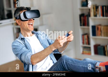 Excited middle aged caucasian man sitting in at the desk and gesturing hands while testing new app via VR device. Cheerful mature man using VR glasses and holds smartphone, virtual reality concept Stock Photo