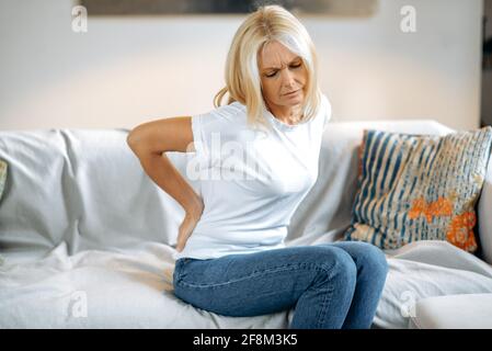 Upset frustrated mature woman suffering from backache, unhappy senior blonde sitting on a sofa at living room, feeling discomfort because of pain in back, have joint problems, need rest. Stock Photo