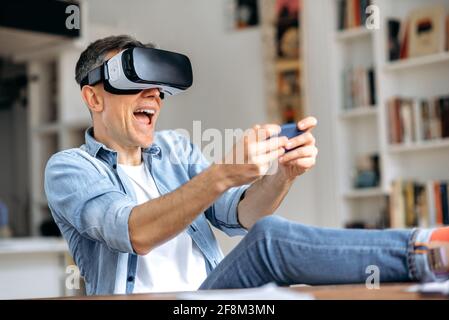 Middle aged caucasian man, freelancer, wearing virtual reality goggles during working break. Excited man testing VR glasses while sits at the desk, smartphone using with VR headset, virtual reality Stock Photo