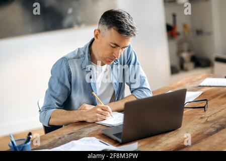 Concentrated adult gray haired caucasian businessman or freelancer, sitting at workplace, taking notes in notebook, studying or working online, using laptop Stock Photo