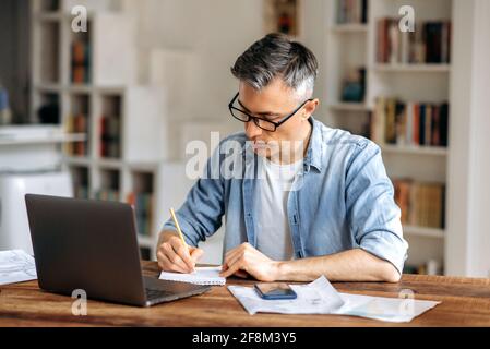 Focused mature gray haired intelligent caucasian businessman or freelancer with glasses, sitting at workplace, taking notes in notebook, studying or working online, using laptop Stock Photo