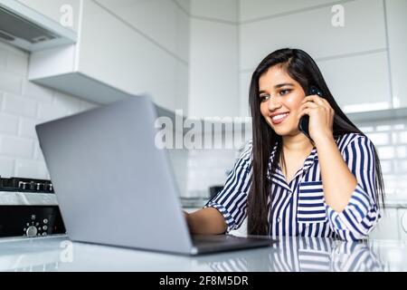 Happy young indian woman at home, talking on mobile phone and using laptop Stock Photo