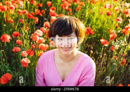 Close up portrait of happy laughing woman with closed eyes in a pink dress relaxing in red poppies flowers meadow in sunset light. A simple pleasure Stock Photo