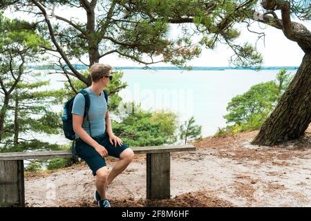 Man with backpack sitting on the bench and enjoying sea view during hiking walk in summer forest. Brownsea Island, England. Local Traveling and relax Stock Photo