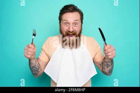 Man with tattoos is ready to eat with cutlery in hand. cyan background Stock Photo
