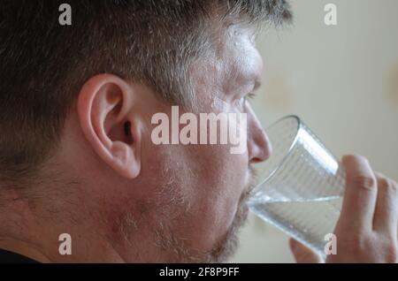 A man with a beard and mustache drinks water from a glass. An adult man holds a transparent glass in his hand. Rear view from the side. Indoors. Lifes Stock Photo