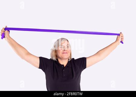 Fit senior blonde woman exercising with mauve resistance band against gym, smiling looking into distance on gray background with copy space. Stock Photo