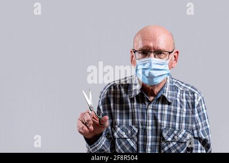 Old bald man with glasses and medical mask with steel scissors in his hand. Copy space. Front view, indoors horizontal shot. Stock Photo