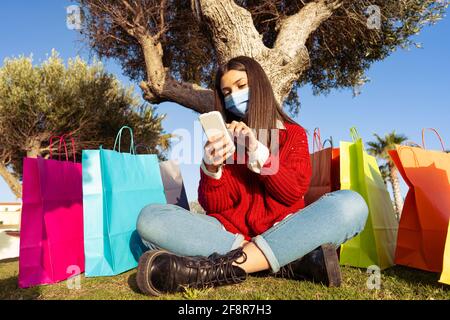 Teenager sitting under tree among colored shopping bags wearing Covid-19 medical protection mask using smartphone searching deals online to buy. New n Stock Photo