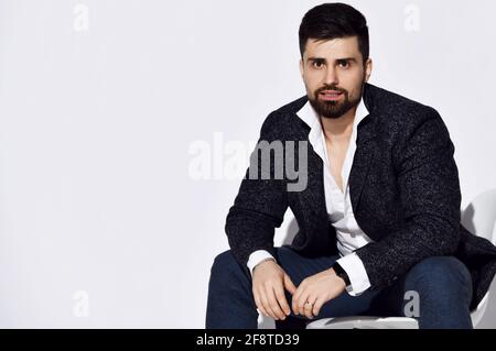 Young stylish bearded man in jeans and casual tweed jacket is sits on chair leaning forward during an interview talking Stock Photo
