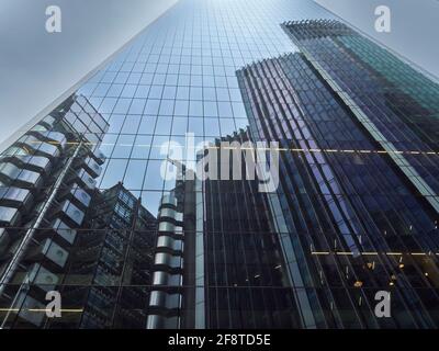 A group of imposing, cyclopean, steel-and-glass London skyscrapers, reflected in the mirrored bulk of one of their fellows, against a grounded sky. Stock Photo