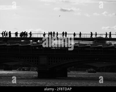 Blackfriars Rail Bridge and the Millennium Footbridge with people walking and enjoying the scenery, all in silhouette, above a choppy Thames. Stock Photo
