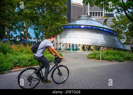 Ride a bicycle in Battery Park urban garden, Lower Manhattan, New York City, New York, USA, North America.  At the back SeaGlass carousel. Stock Photo