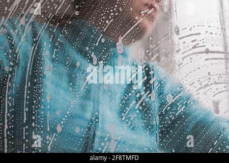 A woman in blue clothes washes a window in a house. Soap foam on the glass. Close-up Stock Photo