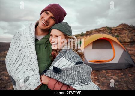Beautiful woman laughing while hugging man wrapped in a blanket during camping at mountain and looking at view Stock Photo